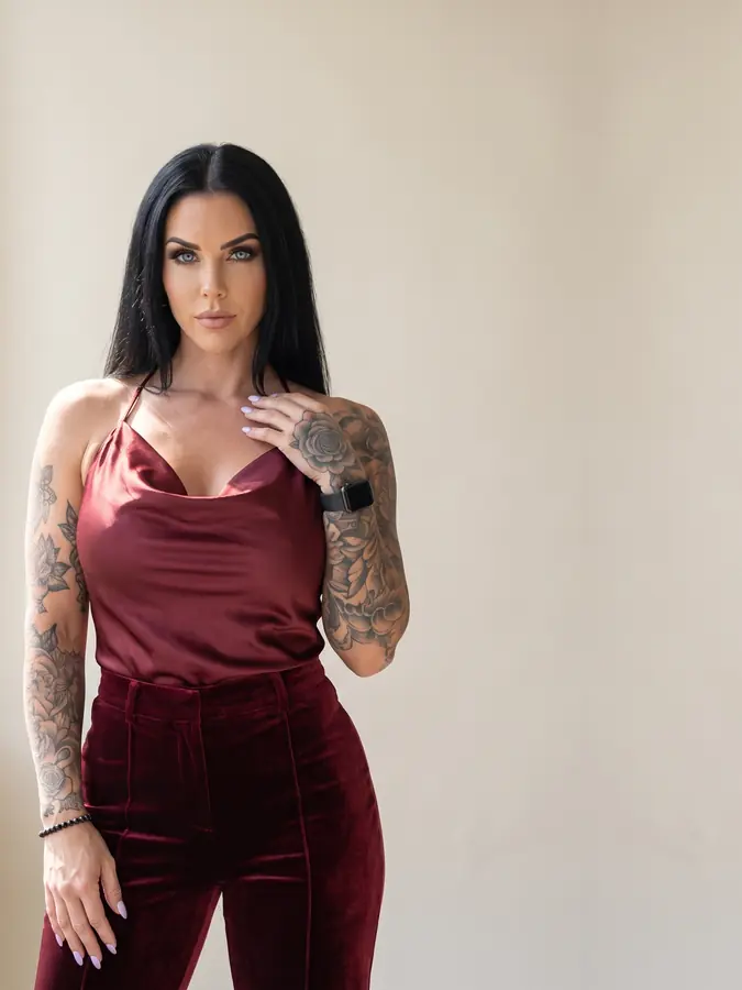 Dahlia Ferrante in an oxblood satin halter top and velvet trousers, one hand at her collarbone, against a pale stone backdrop