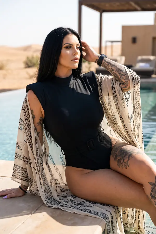 Woman in a belted black high-neck swimsuit and long printed chiffon kimono sitting at the edge of a desert pool