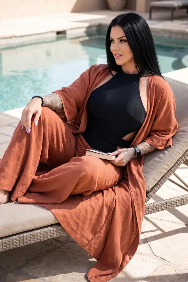 Woman in a black belted halter swimsuit, rust linen kimono and rust wide-leg trousers resting on a lounger by a pool