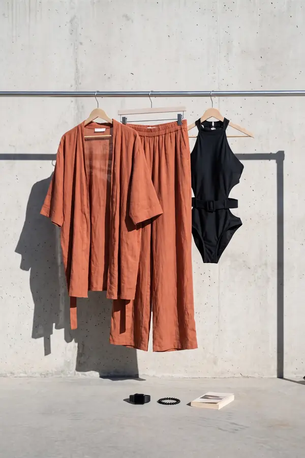 Woman in a black belted halter swimsuit, rust linen kimono and rust wide-leg trousers resting on a lounger by a pool