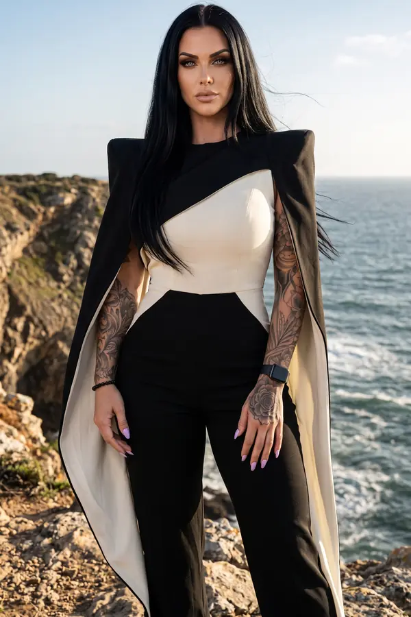 Woman in a black and bone colour-block jumpsuit with a long cape and sharp shoulders standing on a clifftop above the sea