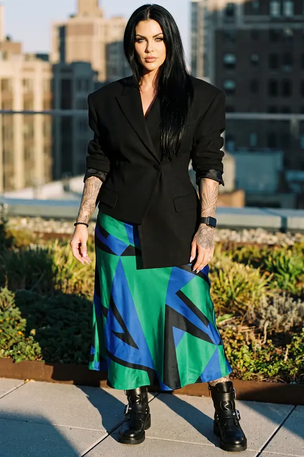 Woman in a boxy black blazer, geometric green and cobalt satin midi skirt and buckled combat boots in a rooftop garden