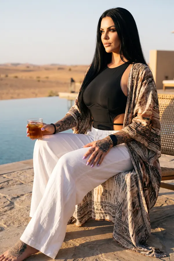 Woman in a black cut-out bodysuit, white linen trousers and a marbled-print kimono seated by a desert infinity pool