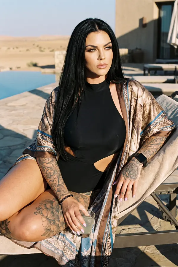 Woman seated on a pool lounger in a black high-neck cut-out swimsuit and a long printed satin kimono, desert behind
