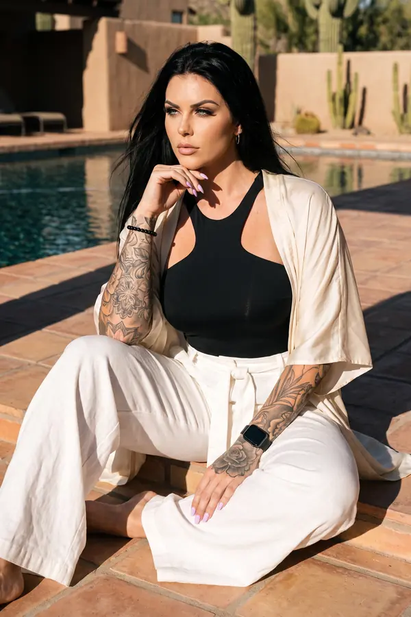 Woman in a black halter bodysuit, ivory satin kimono and ivory linen trousers sitting on terracotta tiles by a desert pool