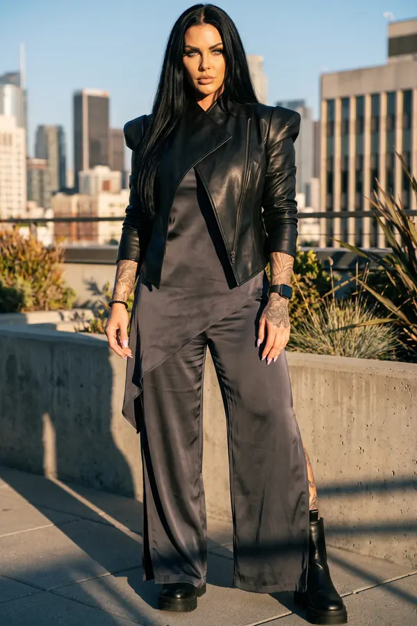 Woman in a cropped black leather moto jacket, charcoal satin top and wide-leg trousers on a rooftop with a city skyline
