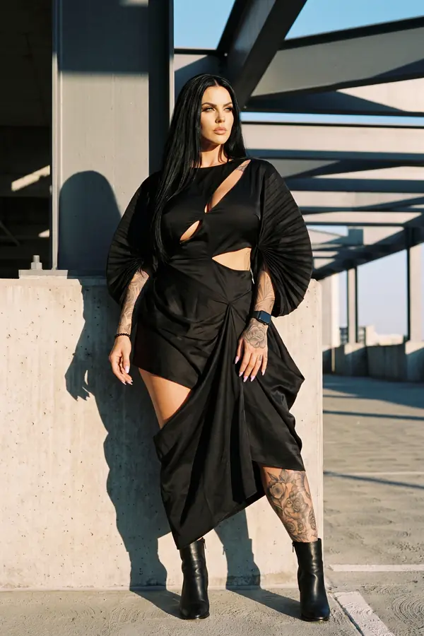 Woman in a black satin cut-out dress with pleated fan sleeves and black ankle boots against a concrete wall
