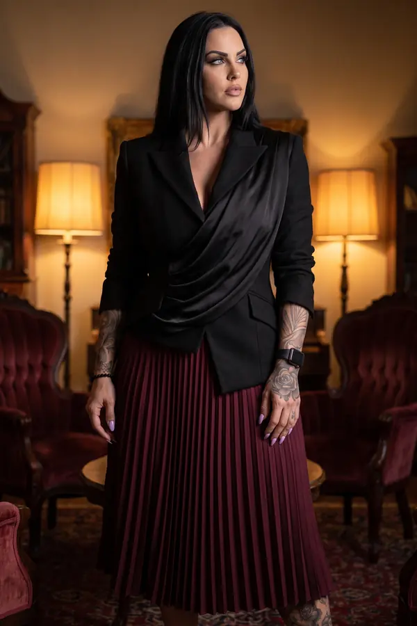 Woman in a fitted black blazer with a satin drape and a burgundy pleated midi skirt standing in a lamp-lit salon