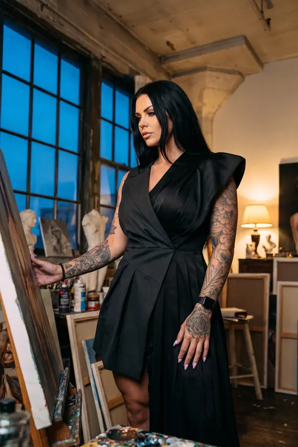 Woman in a black sleeveless blazer dress with winged shoulders and a pleated satin panel beside an easel in a studio at dusk