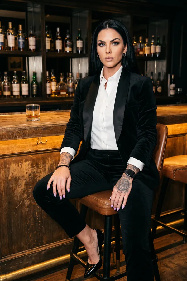 Woman in a black velvet tuxedo blazer with satin lapels and a white shirt seated on a bar stool at a whisky bar
