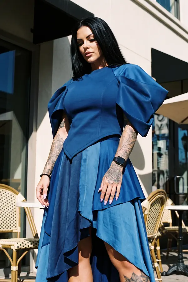 Woman in a blue puff-sleeve top and layered navy satin high-low skirt outside a sunny cafe terrace