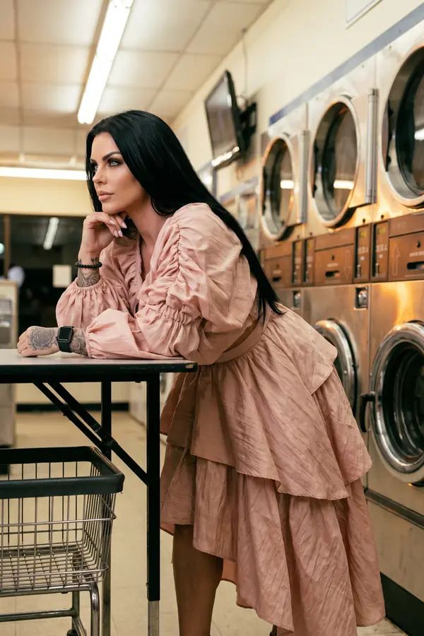 Woman in a blush crinkled satin tiered dress with ruched balloon sleeves leaning on a table in a laundromat