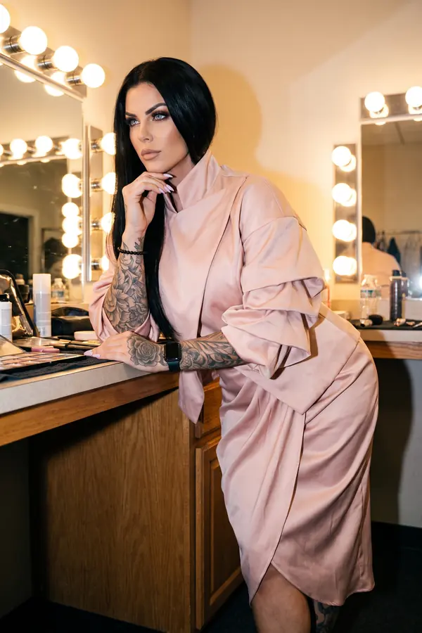 Woman in a blush satin funnel-neck dress with ruched sleeves leaning on a dressing-room counter lit by mirror bulbs