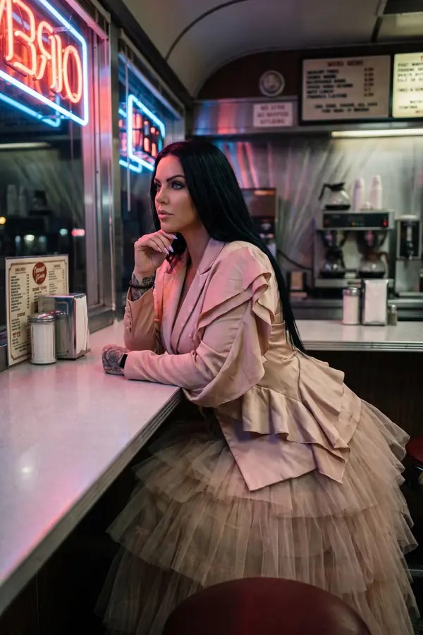Woman leaning on a diner counter at night in a blush satin ruffle blazer and tiered tulle skirt under neon light