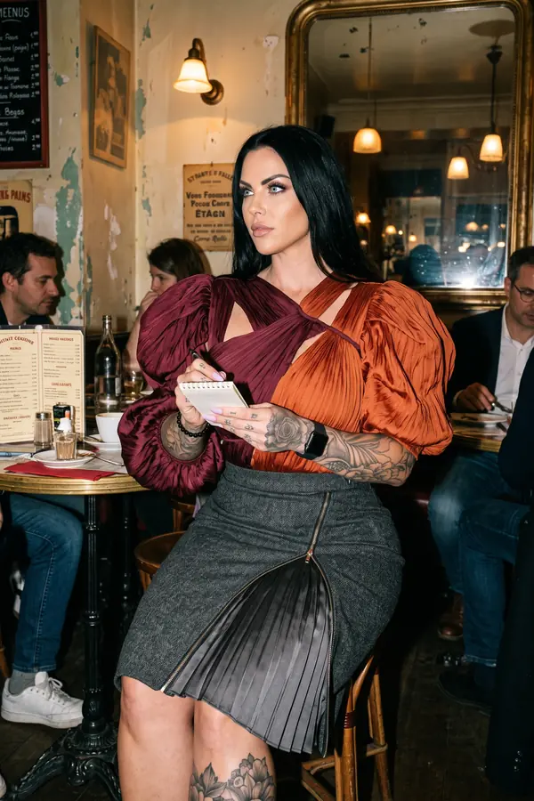Woman in a burgundy and rust pleated satin puff-sleeve top and charcoal tweed zip pencil skirt seated in a brasserie