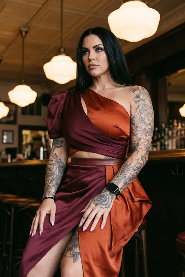 Woman seated on a bar stool in a burgundy and rust satin one-shoulder top and wrap skirt with a tan leather belt
