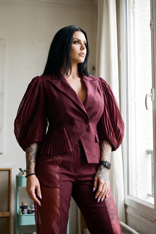 Woman in a burgundy double-breasted blazer with pleated chiffon balloon sleeves and matching trousers by a tall window