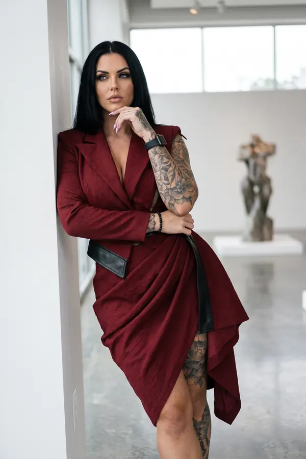 Woman in a burgundy crinkled blazer dress with black leather panels leaning on a white wall in an art gallery