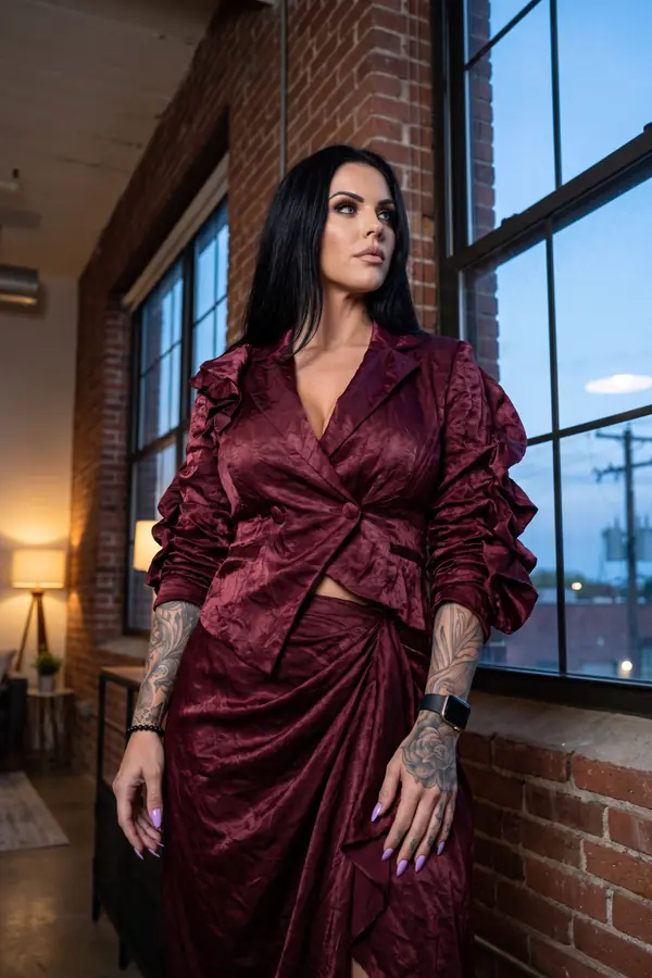Woman in a cropped burgundy crinkled satin blazer with ruffled sleeves and a matching draped wrap skirt in a brick loft