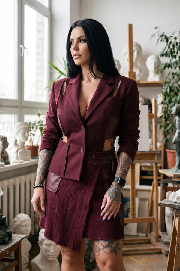 Woman in a burgundy cut-out wool blazer and tweed wrap mini skirt standing among plaster busts in a sculpture studio