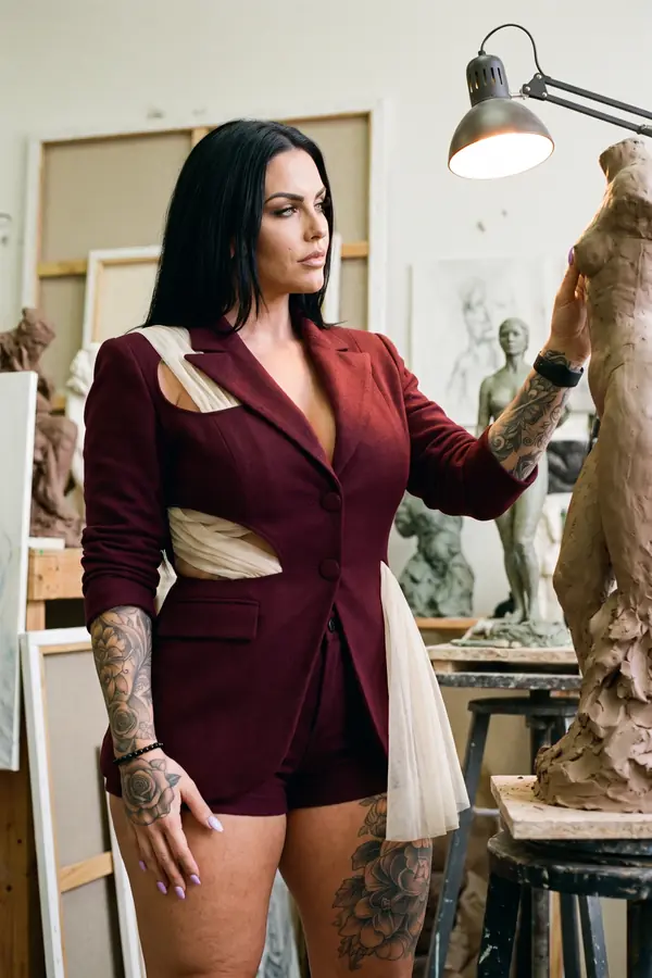 Woman in a burgundy blazer with cut-outs threaded with cream tulle and matching tailored shorts beside a clay sculpture
