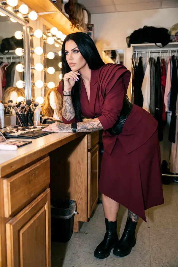 Woman in a burgundy draped blazer, black leather corset belt and asymmetric skirt leaning on a dressing-room counter