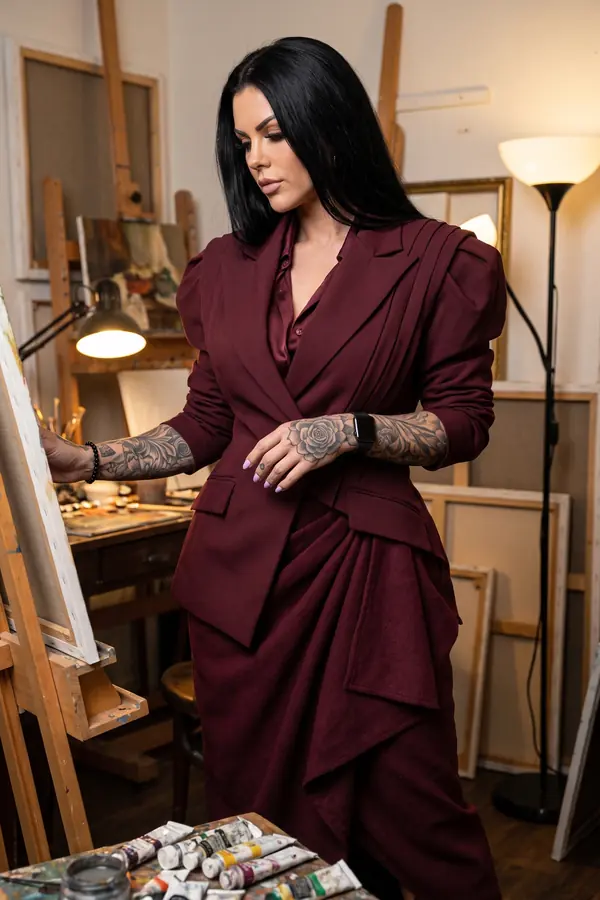 Woman with long black hair in a burgundy draped wool blazer, satin shirt and draped skirt at an easel in a painting studio