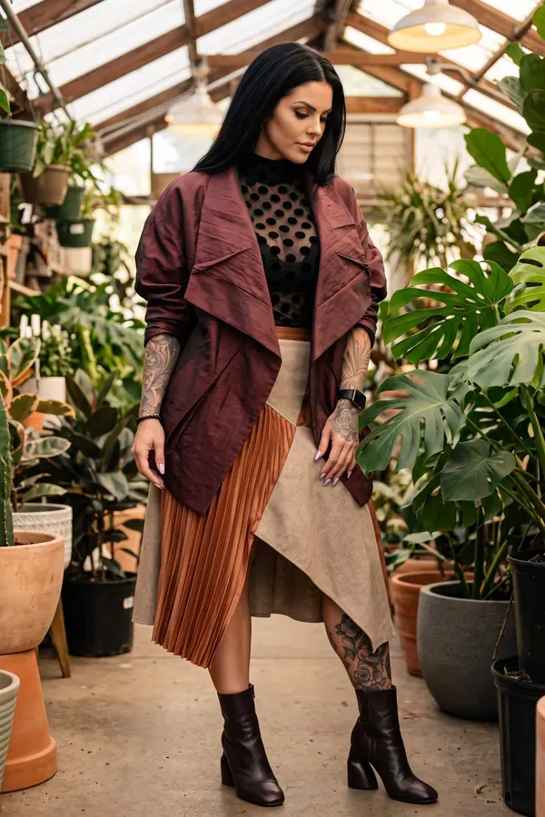 Woman in a burgundy draped-collar jacket, dot mesh top and patchwork pleated midi skirt among greenhouse plants