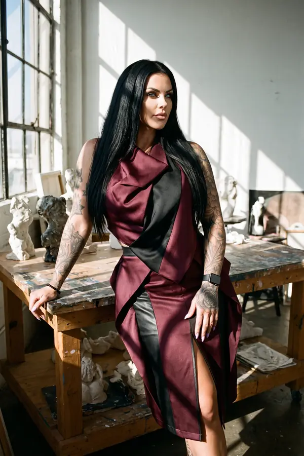 Woman in a sleeveless burgundy satin dress with a black sash panel and leather skirt panel leaning on a sculptor's table