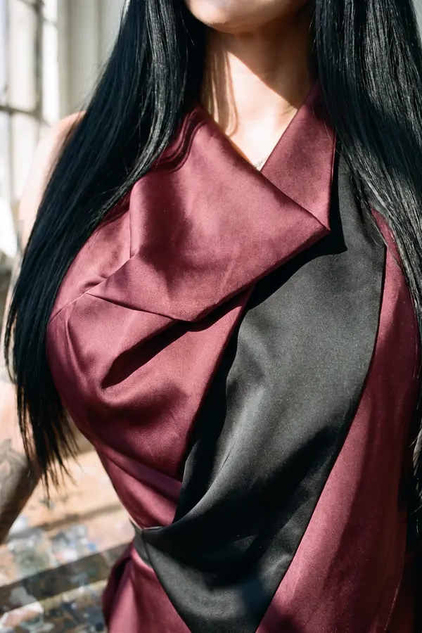 Woman in a sleeveless burgundy satin dress with a black sash panel and leather skirt panel leaning on a sculptor's table