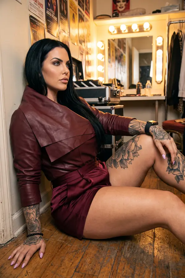 Woman seated on a dressing-room floor in a cropped burgundy leather jacket with a wide folded collar and satin shorts