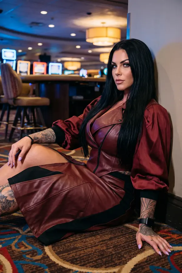 Woman in a burgundy leather zip dress with satin balloon sleeves seated on a patterned casino carpet against a wall