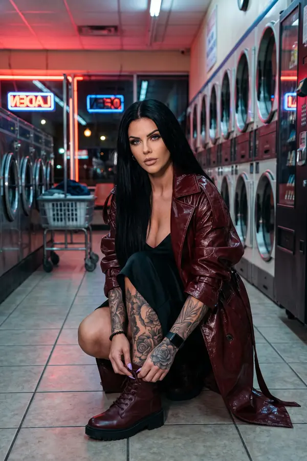 Woman crouching in a laundromat in a glossy burgundy patent trench over a black slip dress, lacing up burgundy lug-sole boots