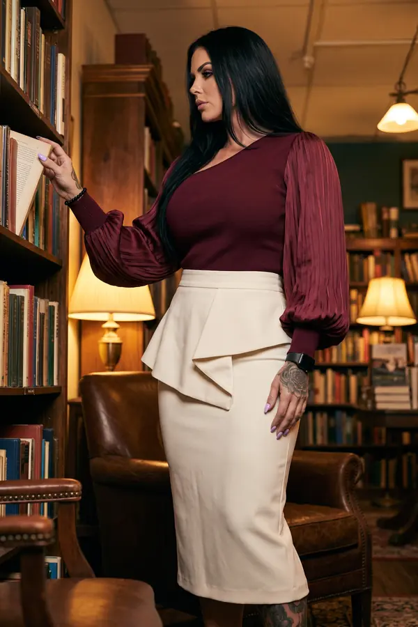 Woman in a burgundy ribbed-knit top with pleated satin bishop sleeves and an ivory peplum pencil skirt in a bookshop
