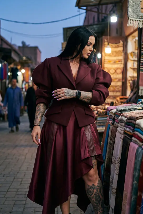 Woman in a burgundy puff-sleeve wool blazer and satin handkerchief skirt walking through a market street at dusk