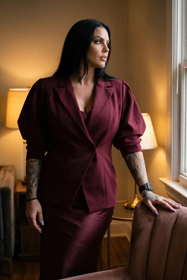 Woman in a burgundy puff-sleeve wool blazer over a burgundy satin slip dress beside a window in a lamp-lit room