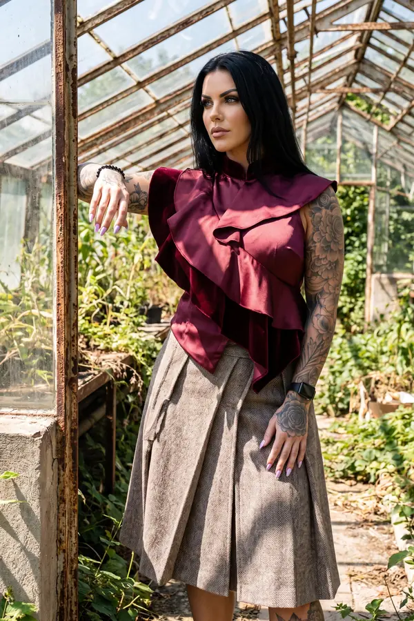 Woman in a burgundy ruffled satin and velvet top and a taupe tweed pleated midi skirt leaning in an old greenhouse