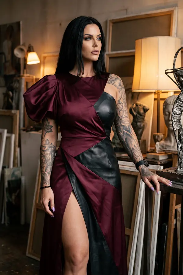 Woman in a floor-length burgundy satin and black leather gown with one puff sleeve standing in an artist's studio