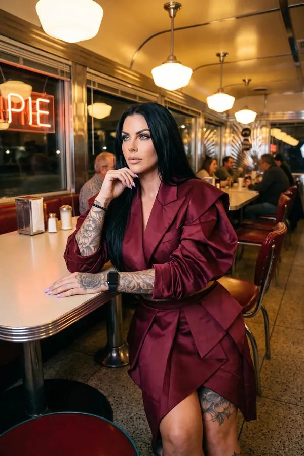 Woman in a burgundy satin blazer with ruched sleeves and a folded-panel mini skirt, seated at a diner table at night