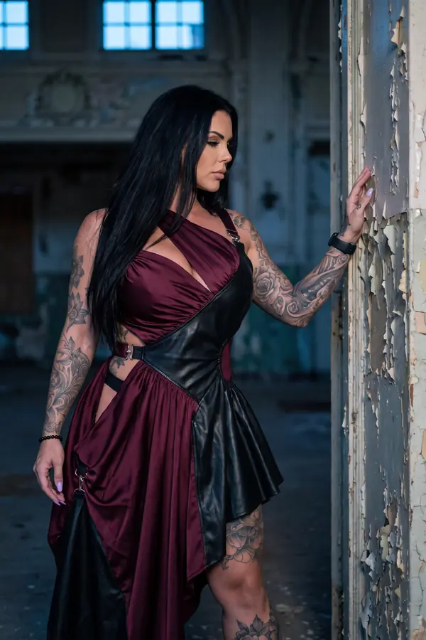 Woman in a burgundy satin halter dress with black leather panels and silver buckles against a wall of peeling paint