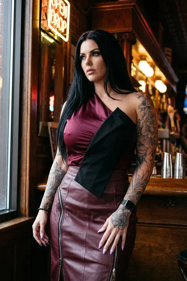 Woman in a burgundy satin one-shoulder top with a black panel and a burgundy leather zip pencil skirt in a wood-panelled bar