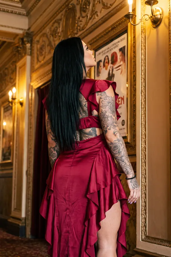 Woman seen from behind in a burgundy satin dress with an open back and cascading ruffles in a gilded theatre corridor