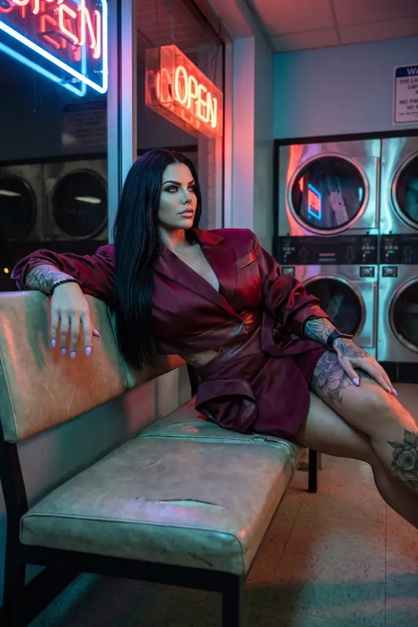 Woman seated on a laundromat bench at night in a burgundy satin wrap blazer with leather trim and burgundy satin mini skirt
