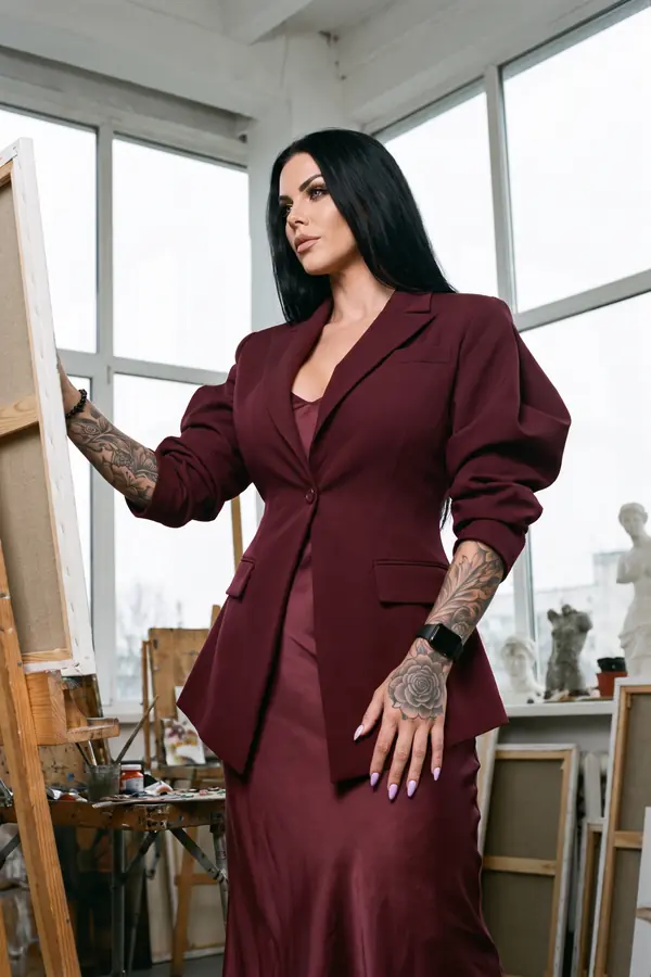 Woman in a burgundy single-button blazer over a burgundy satin slip dress standing at an easel in a painter's studio