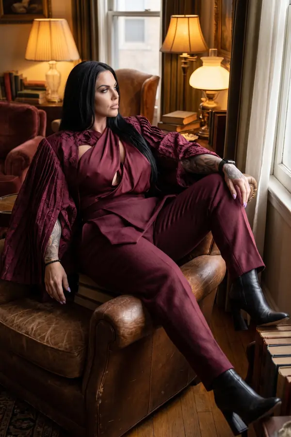 Woman in a burgundy satin twist top, velvet cape jacket and wool trousers seated sideways in a leather armchair by a window