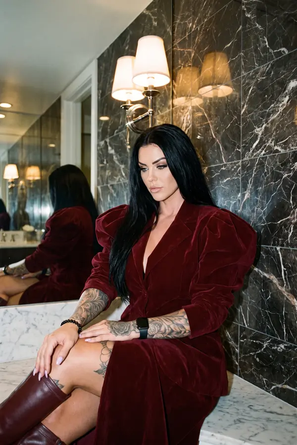 Woman in a burgundy velvet blazer dress with puffed shoulders and burgundy knee boots seated in a marble hotel bathroom