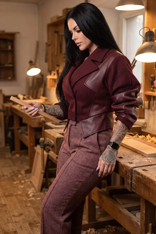 Woman in a cropped burgundy wool and leather jacket and tweed trousers leaning on a bench in a carpentry workshop