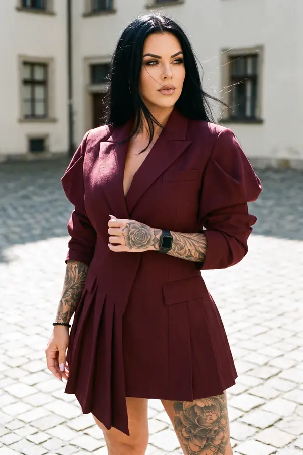 Woman in a burgundy wool blazer dress with an asymmetric pleated panel standing on cobblestones in an old courtyard