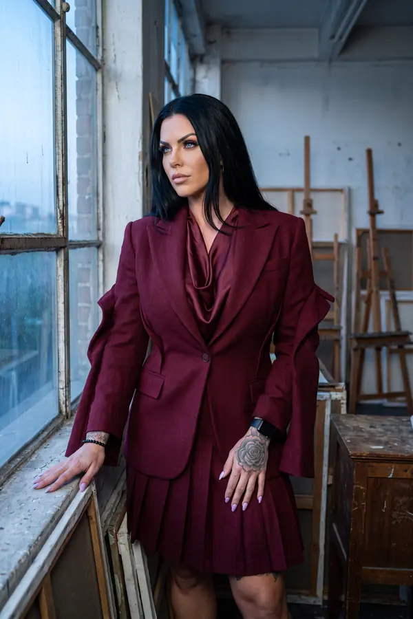 Woman in a burgundy wool blazer with split sleeves, a satin cowl-neck blouse and a pleated mini skirt by a studio window