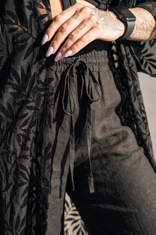 Woman in a black cage cut-out swimsuit, black burnout kimono and black linen trousers sitting by a desert pool at golden hour
